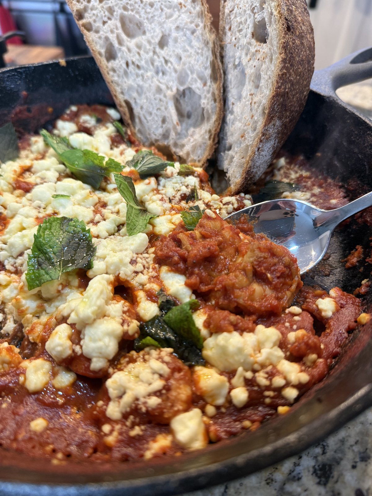 Spiced shrimp saganaki with tomato and feta in a cast-iron skillet, topped with torn mint, with crusty bread alongside